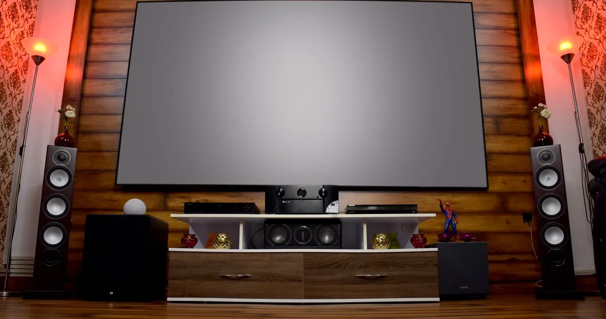 Sala de home theater com tela grande e caixas de som ao lado de um móvel de madeira, ilustrando a escolha entre barra de som ou caixas